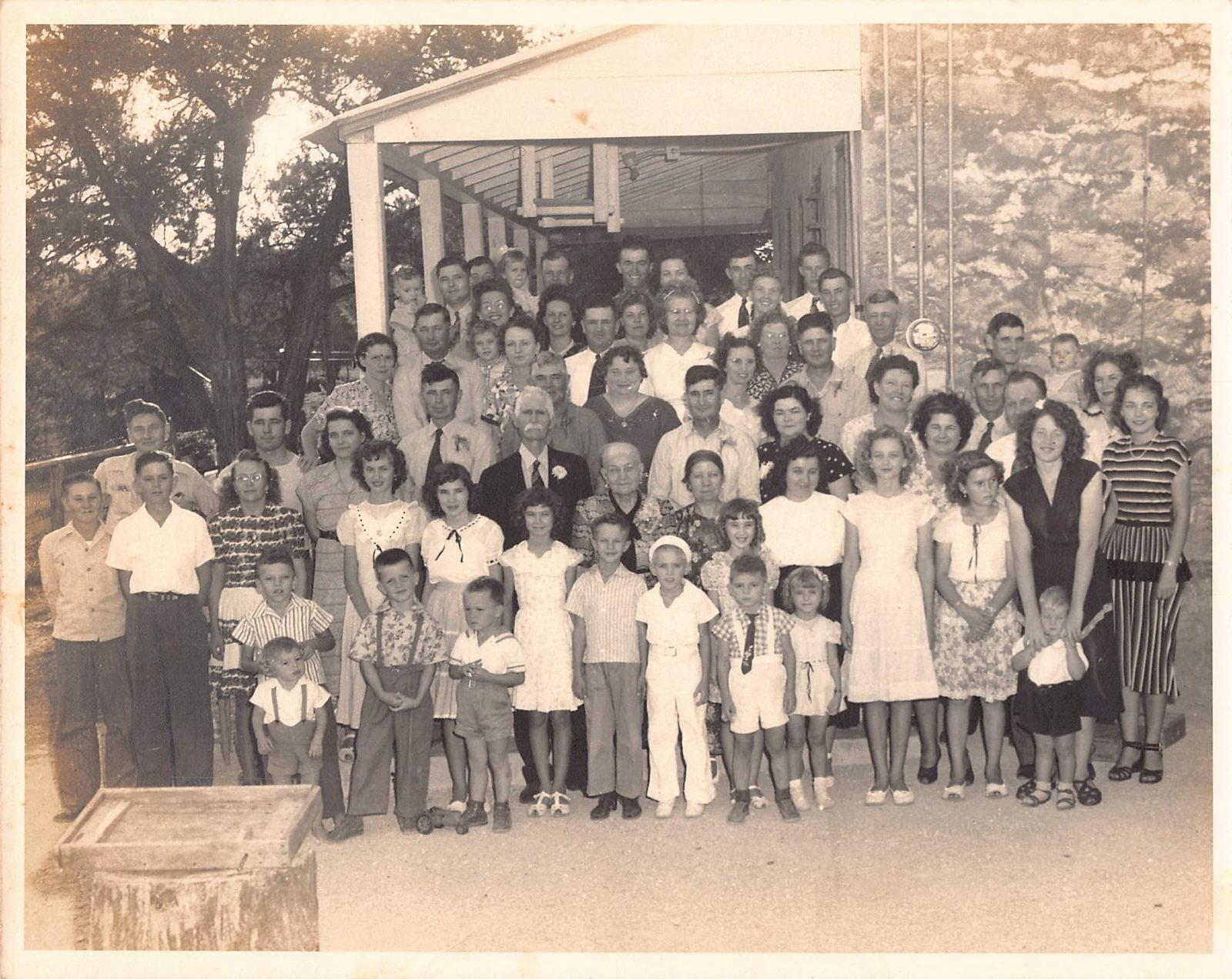 Around eighty members of the Behrends family posed in front of a stone building with a covered porch, in clothing of the late 1940s or early 1950s.